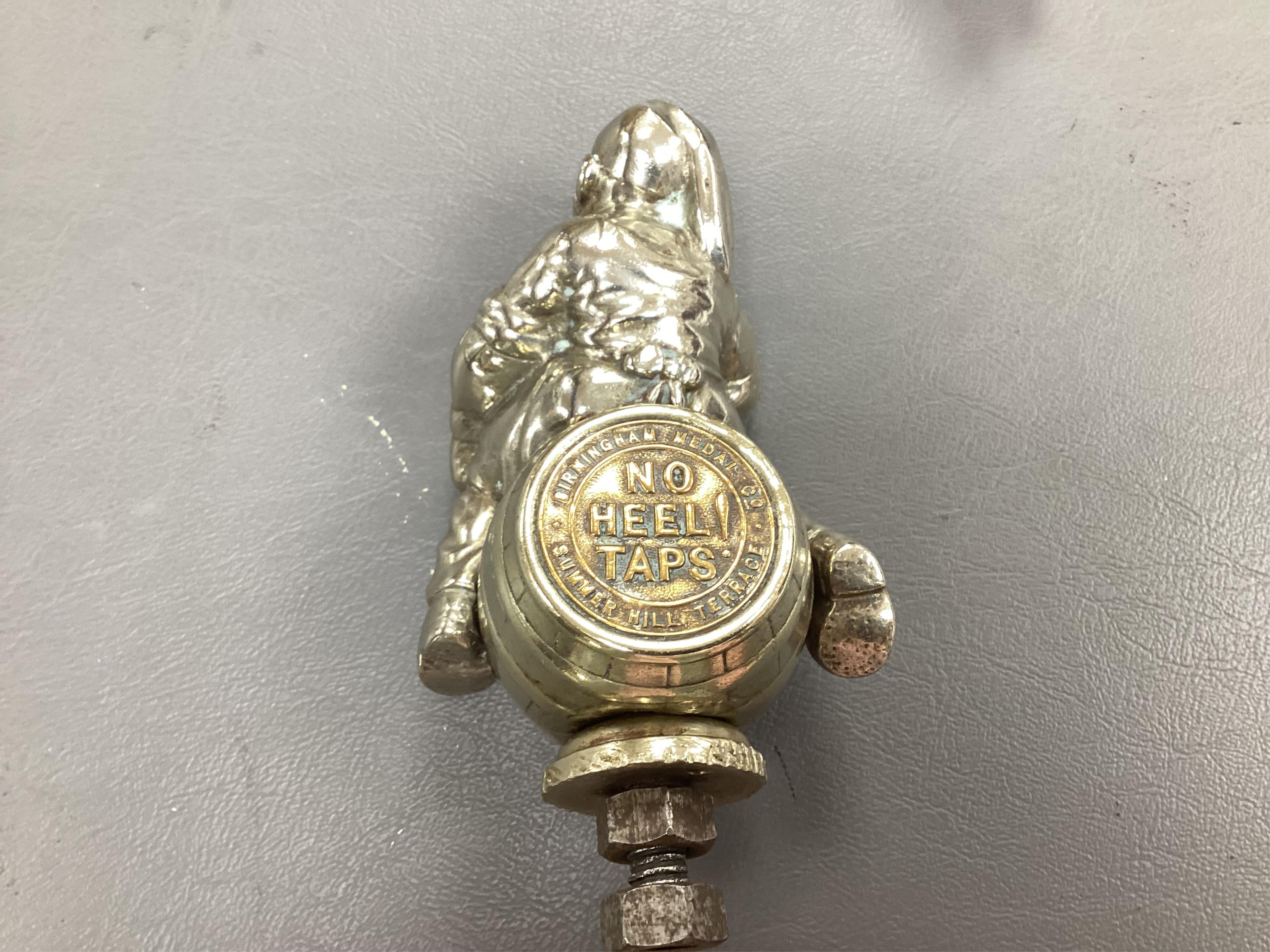 Seven chrome and brass car mascots, including a Mercedes-Benz logo, a figure of a blacksmith, a caricature figure on a beer barrel, a golf ball in mid flight, etc. together with a chromed housing unit for an electric lam